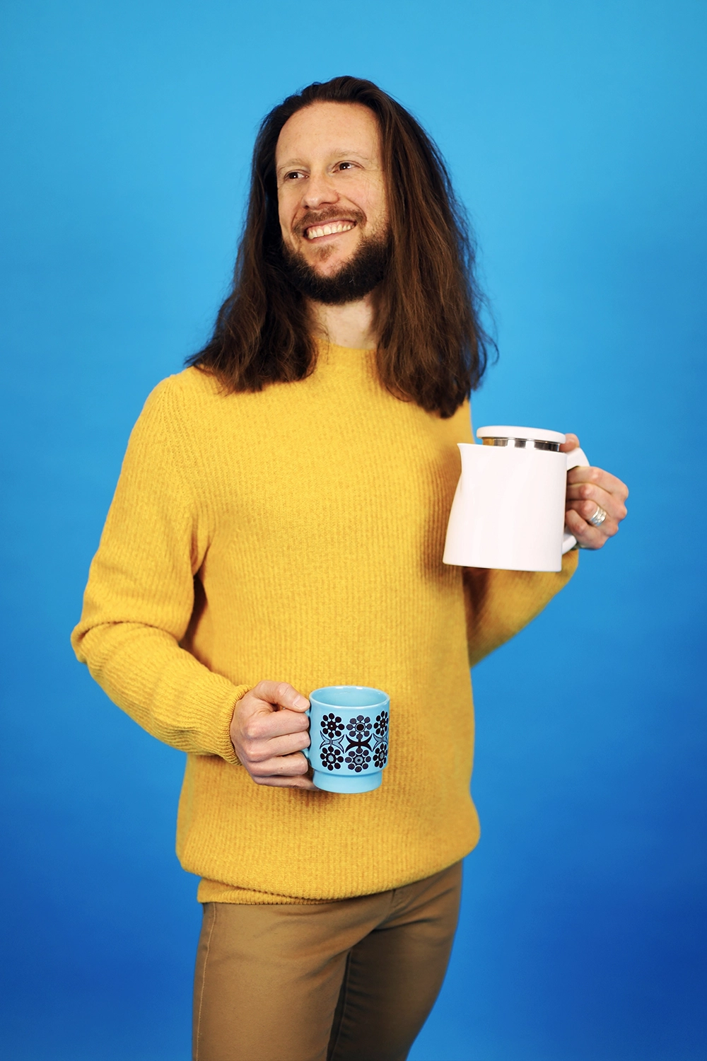 Homme avec cheveux longs souriant, tenant une tasse et une cafetière.