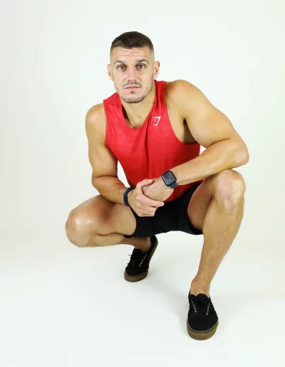Coach sportif accroupi en débardeur rouge et short noir dans un studio photo.