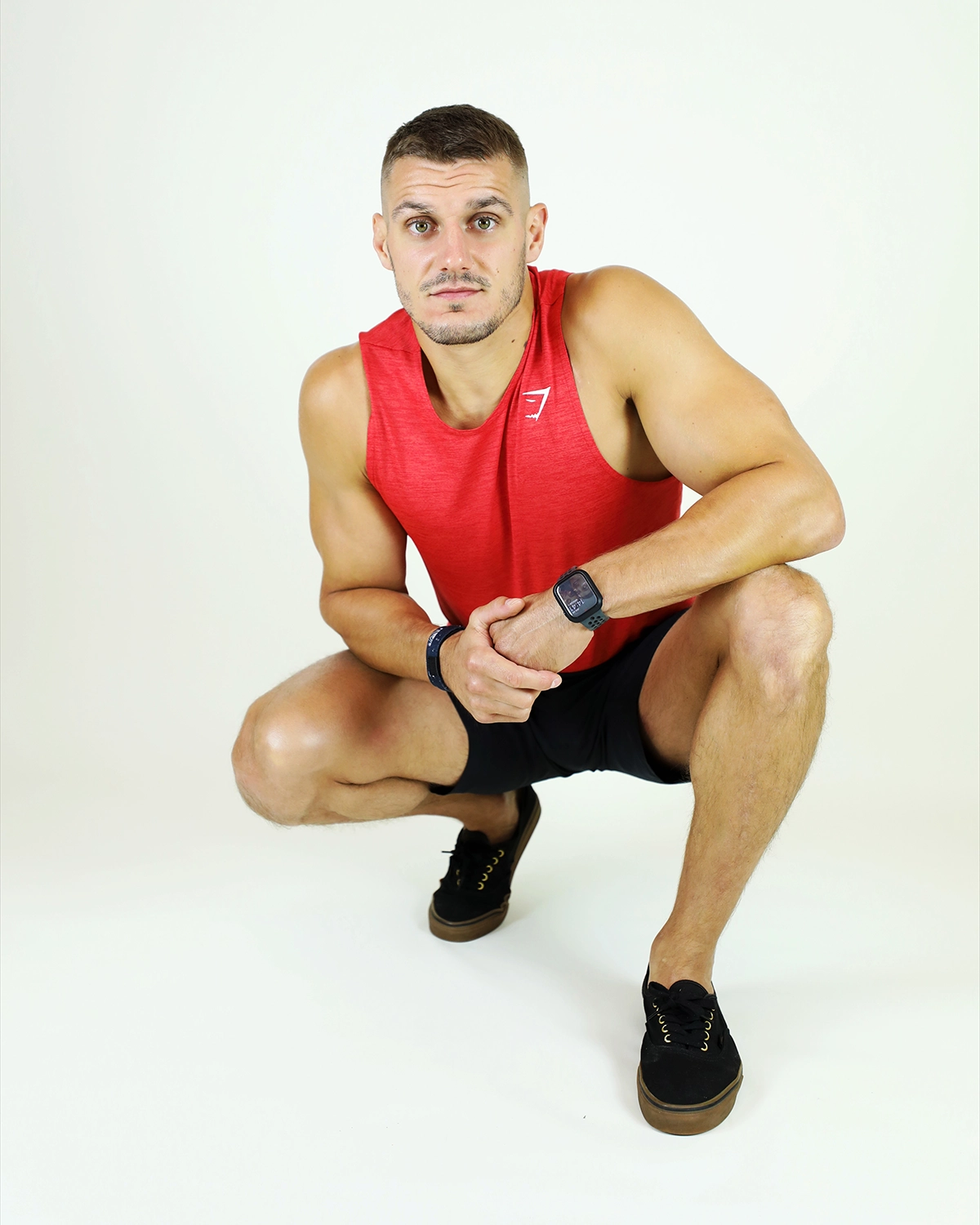 Coach sportif accroupi en débardeur rouge et short noir dans un studio photo.
