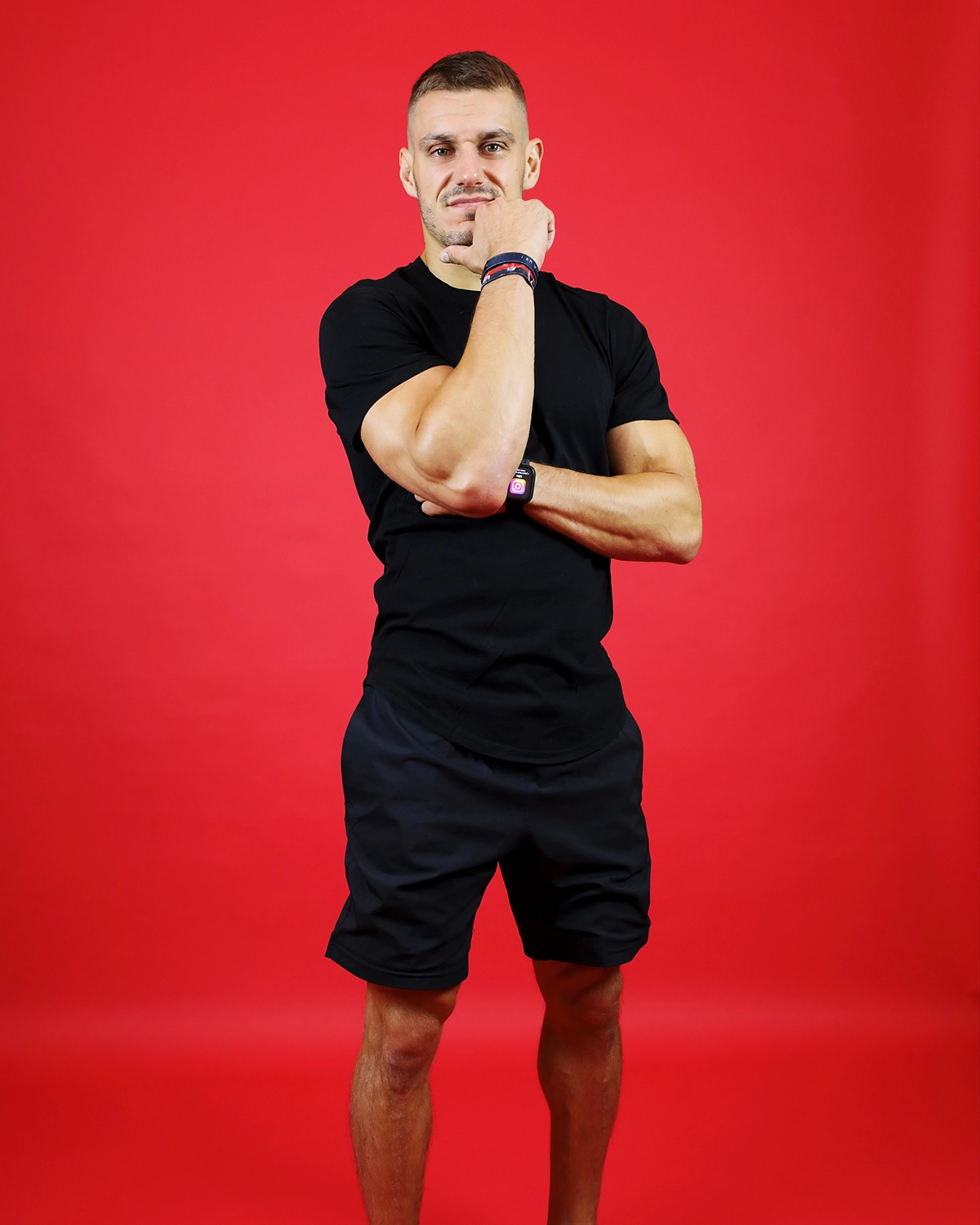 Coach sportif en position décontractée, portant un t-shirt noir et un short sur fond rouge.