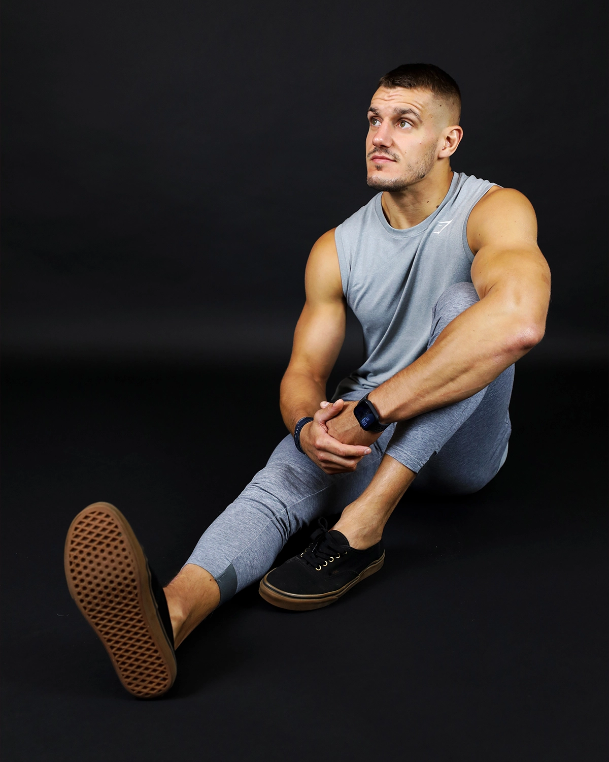 Portrait studio d'un coach sportif assis en position décontractée, portant un débardeur gris.