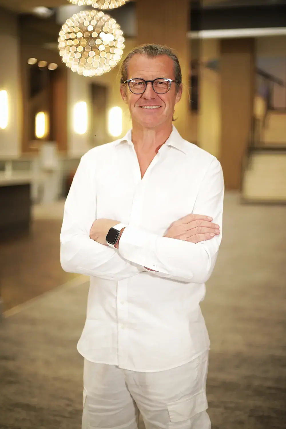 Portrait professionnel en intérieur lumineux Homme en chemise blanche, bras croisés, photographié dans un intérieur lumineux avec des suspensions en arrière-plan.