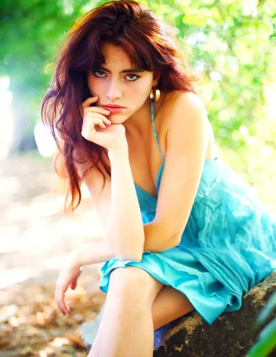 Femme aux cheveux roux vêtue d’une robe turquoise, assise dans un environnement végétal lumineux.