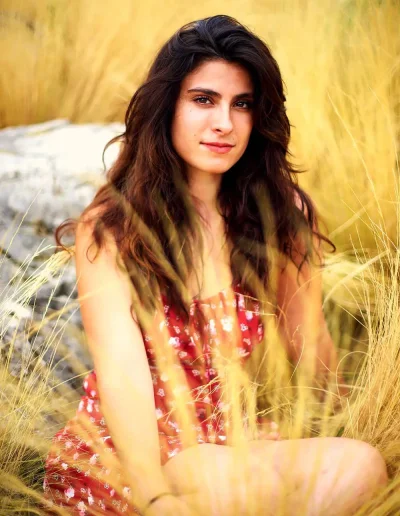 Femme brune assise dans des hautes herbes dorées, vêtue d’une robe rouge à motifs.