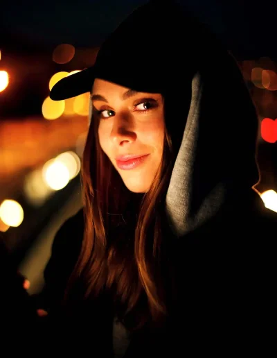 Femme portant une casquette et une capuche noire, photographiée de nuit devant des lumières floues.