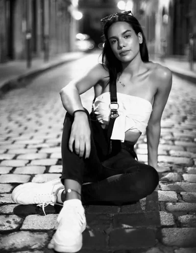 Femme assise dans une rue pavée la nuit, photographiée en noir et blanc.