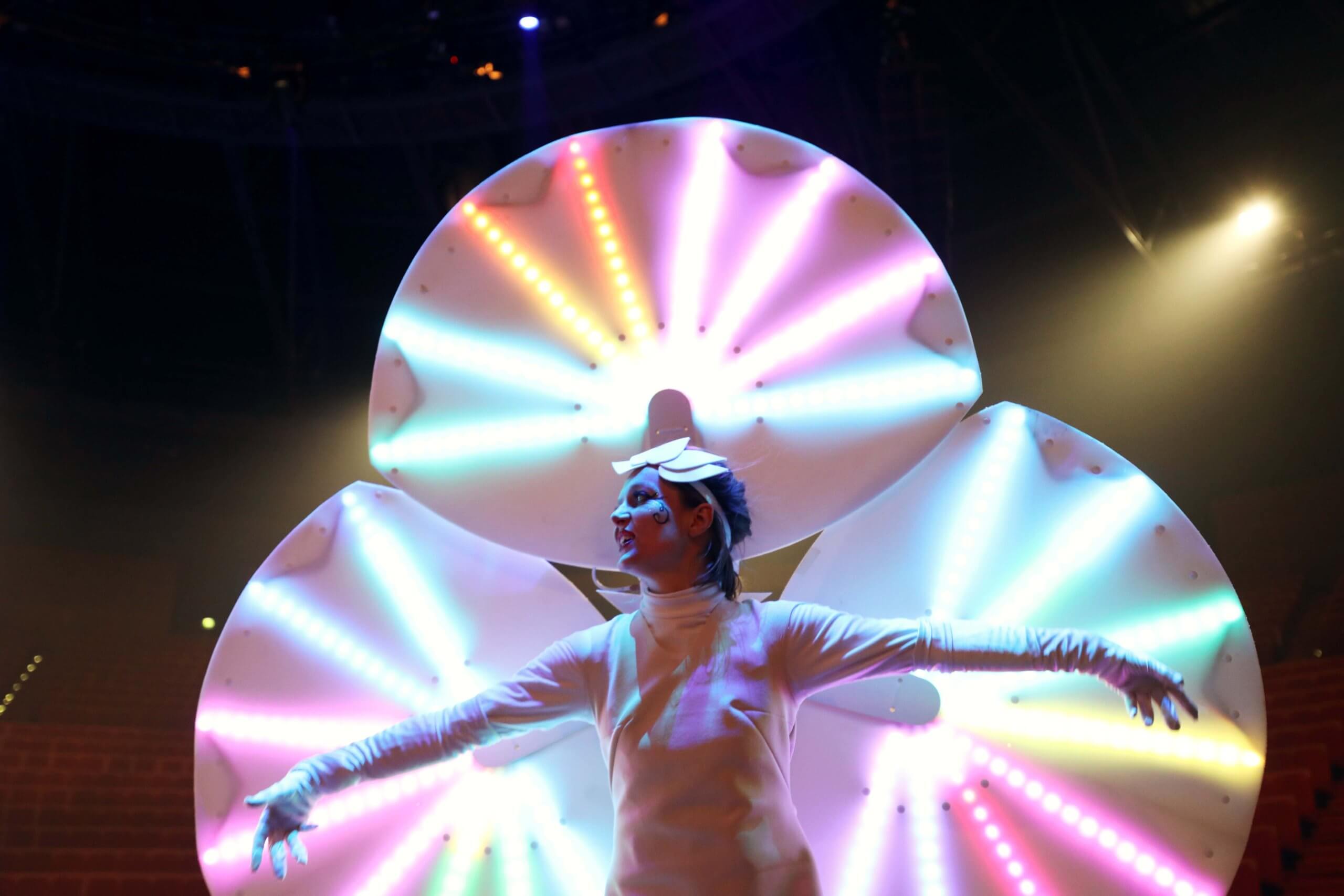 Artiste en costume blanc - spectacle lumineux Un artiste en costume blanc ouvre les bras devant de grands éléments circulaires éclairés de couleurs.