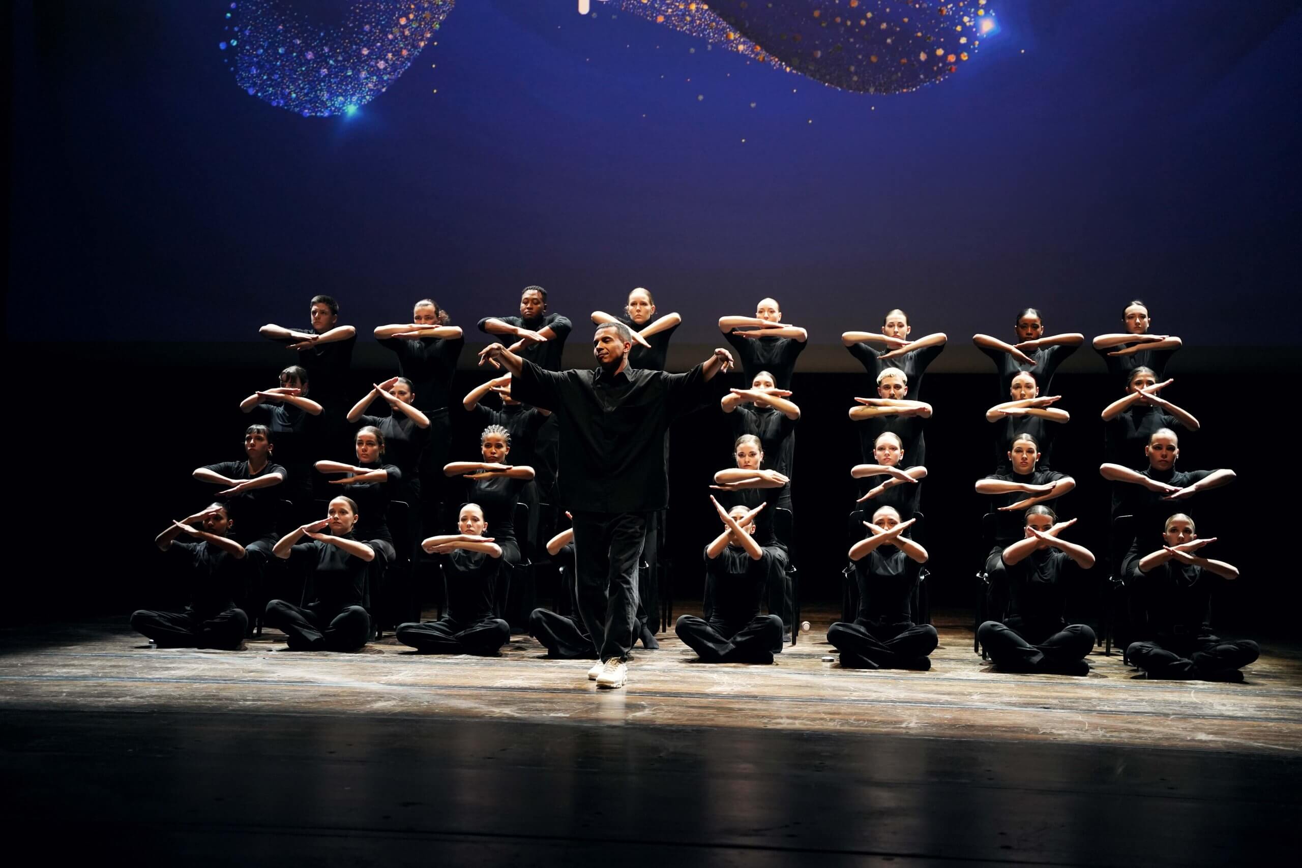 Spectacle de danse Un ensemble d’artistes vêtus de noir réalise des gestes synchronisés autour d’un interprète au centre de la scène.