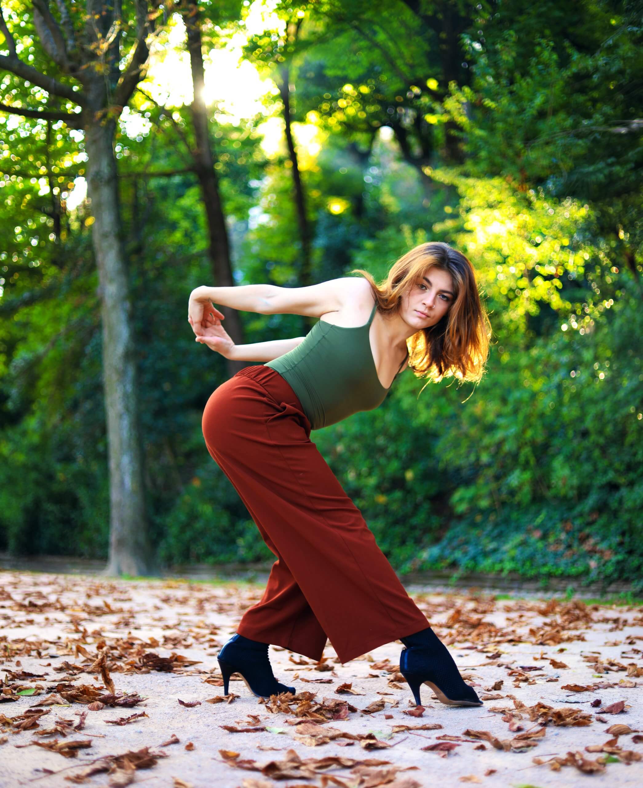 Portrait de danseuse en extérieur Danseuse en haut vert et pantalon rouge dans une pose inclinée, devant des arbres à contre-jour.