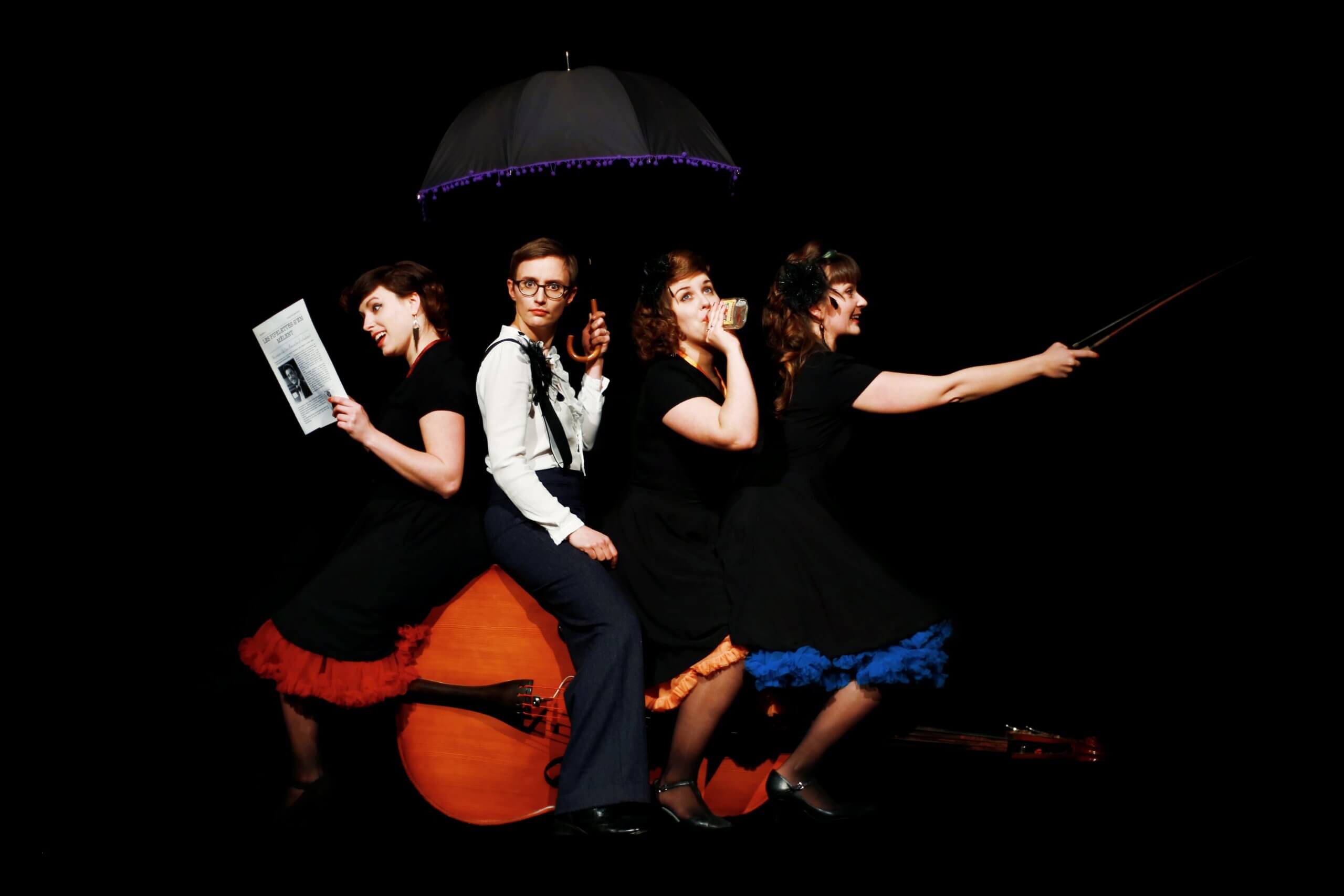 Les Piplettes - photo promotionnelle avec contrebasse Les quatre artistes des Piplettes posent autour d’une contrebasse, avec un parapluie et des accessoires, sur fond noir.