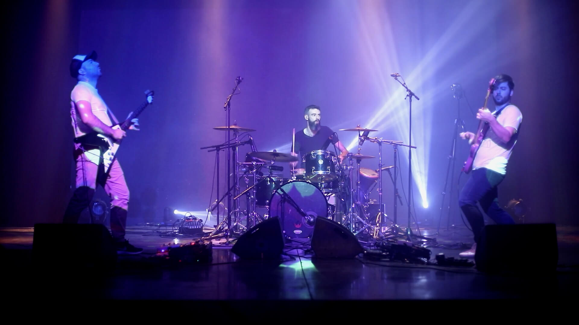 Captation vidéo d’un trio en concert Trois musiciens en concert, avec une batterie au centre de la scène, sous un éclairage violet.