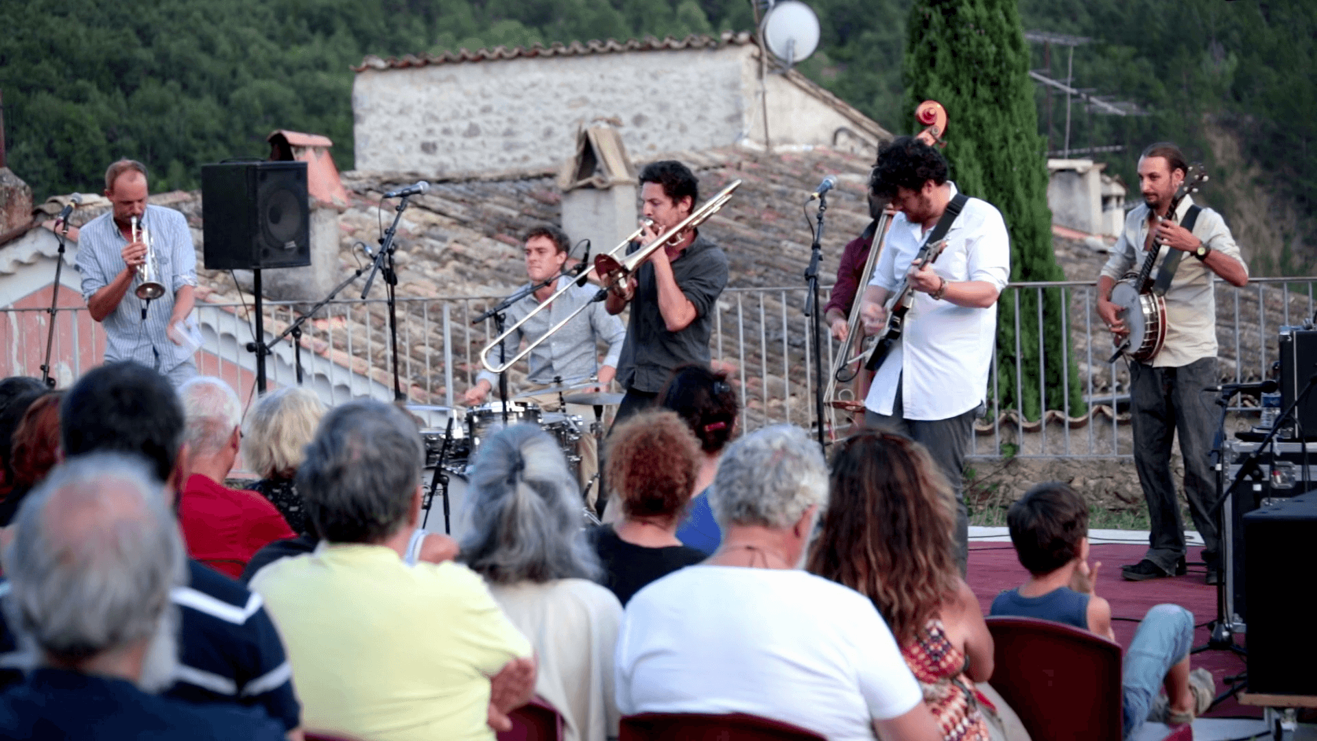 Captation vidéo d’un concert en plein air Musiciens jouant en plein air devant un public assis, avec une trompette, un trombone et un banjo visibles.