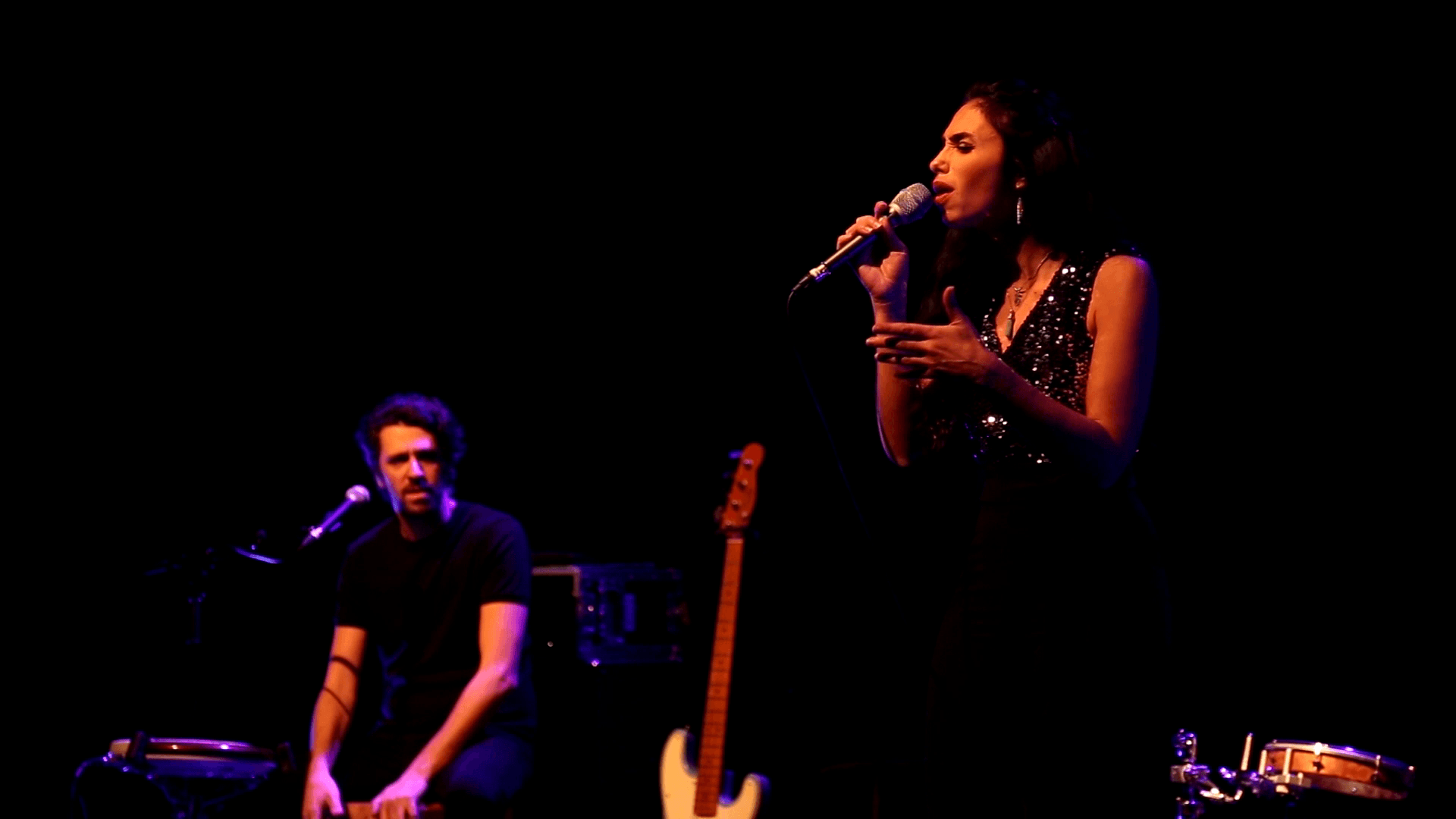 Captation musicale : chanteuse et accompagnateur sur scène Chanteuse au microphone et musicien assis à ses côtés, sur une scène éclairée de lumières chaudes et violettes.